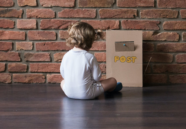 Kind in weißem Body sitzt vor Karton Postkasten